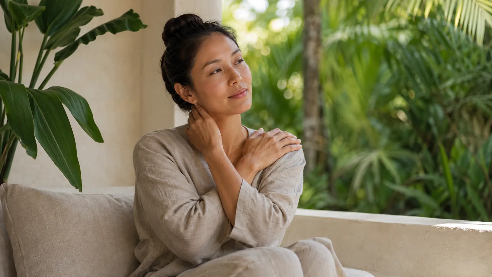 Woman gently holding her neck and shoulder while seated beside tropical greenery